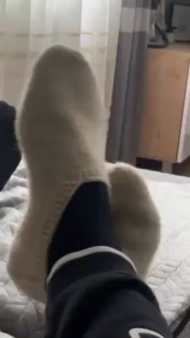 The video shows a close-up of a person's legs and feet resting on a bed. The person is wearing black sweatpants with a white stripe on the side and black socks underneath off-white, knitted slipper socks. The bed has a light gray patterned blanket. In the background, there is a beige curtain, a light-colored dresser, and a blurred object on top of the dresser. The audio suggests a conversation is taking place. The focus is on the footwear and legs.