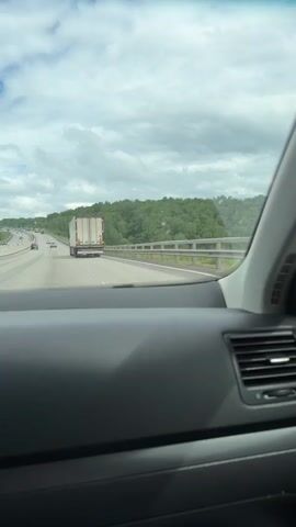 The video provides a first-person perspective from the passenger seat of a car traveling on a multi-lane highway.

Initially, the camera faces forward, showing the road ahead where a white semi-truck with a reddish-brown strip on its cargo container is visible in the middle lane, moving in the same direction. Other distant cars are also on the highway. Lush green forested hills flank both sides of the road, which is separated from the scenery by a silver metal guardrail. The sky above is overcast with thick clouds, indicating a cloudy daytime. The car's dark gray dashboard and a section of the windshield are visible in the foreground.

The camera then slowly pans to the right. The car's dashboard and a passenger-side air vent become more prominent. As the pan continues, the car's side mirror comes into view. The reflection in the side mirror shows a person, presumably the filmer, holding a smartphone, capturing the scene. The reflection also reveals more of the cloudy sky and the green hills.

Throughout the video, upbeat electronic or dance music is playing, audible from inside the car. The scene depicts a journey through a scenic, possibly rural or semi-rural, landscape. Given the context, the location is Sanatoriya Alkino, Russia.
