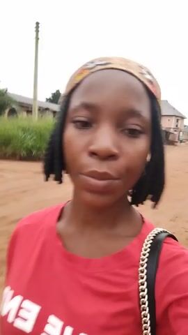 A young woman with braided hair and a bandana walks down a dirt road. She is wearing a red t-shirt and carrying a black purse with a chain strap. The background consists of houses and vegetation.