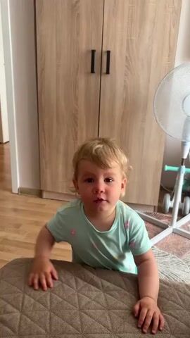 A blonde toddler wearing a light blue t-shirt with a floral pattern is on a beige mat. They are leaning over the mat, with their hands on its surface. The toddler then moves forward, with their head going down towards the mat. Their body follows, and they end up lying on the mat on their stomach, facing away from the camera. In the background, there is a light wood wardrobe with two black handles, a white fan with a light blue base, and a cream-colored rug. The floor is wooden.