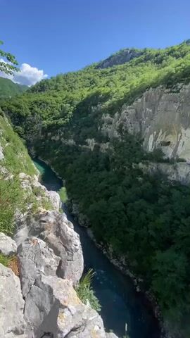 A breathtaking aerial view captures a deep river canyon in Vlahovići, Montenegro, under a clear blue sky with scattered white clouds. The camera slowly pans downwards and slightly to the right, revealing the grandeur of the natural landscape. A vibrant turquoise-blue river flows through the base of the canyon, snaking between towering, steep cliffs. The canyon walls are densely covered with lush green trees and vegetation, contrasting with the exposed, lighter grey rock faces that show geological layering and some small caves or indentations. Large, rugged rocks are visible in the foreground on the left, emphasizing the height and steepness of the viewpoint. The scene is bathed in bright sunlight, highlighting the textures of the rock and the verdant foliage. No people or text are visible in the video, emphasizing the untouched natural beauty of the gorge.