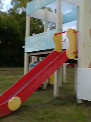 The video appears to be filmed at a playground in Smolino, Russia. The setting is an outdoor play area with various equipment, including a red slide, a sandbox with yellow and red trim, and what looks like a multi-purpose outdoor gym with a blue roof. There are also benches and a wooden fence visible. In the background, there are houses and trees. The time of day appears to be late afternoon or early evening, as suggested by the soft, diffused light and the color of the sky, which is partly cloudy with hints of pink and orange. The weather seems to be mild.

The main subject in the video is a young woman wearing a light brown hooded jumpsuit. She is standing in the playground, looking towards the camera and speaking. Her demeanor appears relaxed and casual. The activity taking place is simply a person in a playground. There is no visible text in the video. The overall mood is calm and perhaps slightly melancholic due to the fading light. The presence of playground equipment suggests a place for recreation, likely in a village or small town setting, given the houses and rural feel of the surrounding area.
