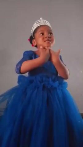 A little girl with a dark complexion and short black hair with small red bows is wearing a silver tiara and a dark blue tulle gown with ruffled details at the waist. She stands in front of a gray wall and moves her hands, forming them into fists and making small gestures as she speaks.