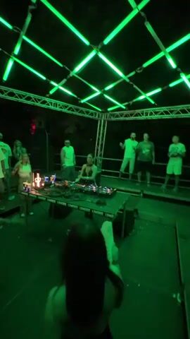 A DJ is playing music on a console in front of a group of people. Green lights are installed on a metal structure above the DJ booth and the surrounding area. The DJ is wearing a black outfit. Some people in the crowd are dancing. The event appears to be taking place outdoors at night.