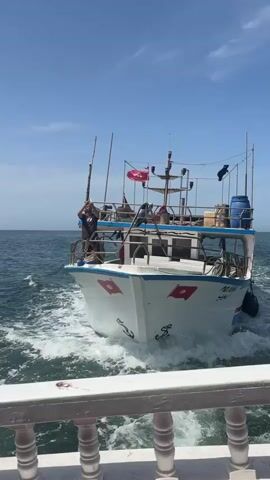 A fishing boat is sailing on the sea under a clear sky. The boat is white with red and blue accents. There are people on the deck of the boat. The boat is moving through the water, creating a wake. The camera is looking from another boat which is in the foreground, partially obscuring the view of the sea and the fishing boat.