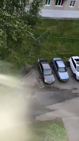The video shows an exterior view of a Soviet-era apartment building in Beloretsk, Russia. The building is a multi-story concrete structure with numerous windows and balconies. Several cars are parked in front of the building, on a paved area surrounded by grass and trees. The sky is overcast, suggesting it may be late afternoon or early evening. The lighting within some of the apartment windows indicates that people are inside their homes. The camera is held at an angle, possibly from another building or a high vantage point, and the footage is somewhat shaky. There are no people visible in the frame, and no text is present. The overall impression is a quiet residential scene in a Russian town.
