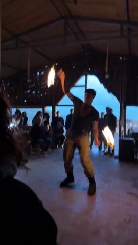 Inside a building with a high, thatched roof, a man performs a fire dance in front of a small audience. He wears a black shirt, khaki pants, and black boots. He holds flaming torches in each hand, skillfully twirling and maneuvering them. The audience, a mix of seated and standing individuals, watches the performance. Some are seated on chairs and appear to be older, while others stand near a large window looking out at the landscape. Light streams through the window, illuminating the space along with the torches. Additional flaming torches stand upright on platforms near the windows. The floor is a rough, light-colored surface. A speaker sits near the back of the room. A person's back and the side of their head is visible in the foreground of the video.