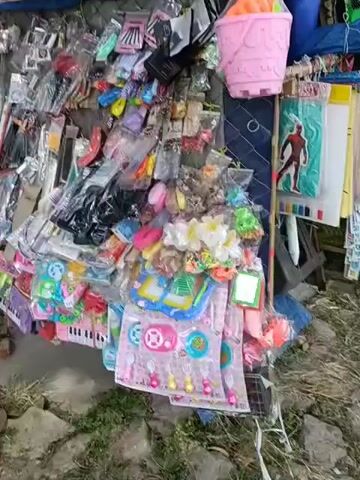 A street vendor is showcasing their wares. The stall is piled high with a colorful assortment of plastic toys and accessories, including miniature keyboards, hair clips, toy telephones, and a pink bucket. Two men are sitting on a ledge in front of the stall, engaged in conversation. One man, wearing a hooded jacket and mask, is looking down at his hands. The other man, wearing a cap and vest, is looking towards the vendor's stall. A motorcycle is parked behind the stall. The scene appears to be outdoors, with a stone path and some greenery visible.
