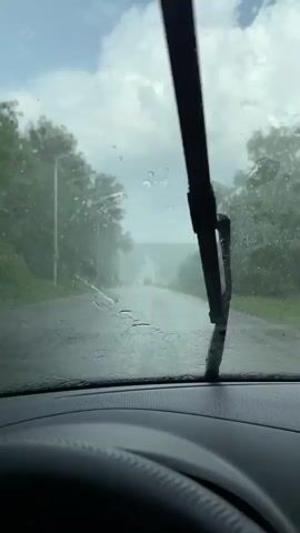 The video begins with a view from inside a car, looking out through the windshield which is covered in raindrops. The road ahead is wet and appears to be a rural highway surrounded by trees on either side. The sky is overcast with clouds. In the distance, there are orange traffic cones and what appears to be a construction barrier on the road. The windshield wipers are activated and are seen clearing the rain from the windshield. The view moves forward as the car travels down the road. The video ends with the wipers having cleared a significant portion of the windshield, revealing more of the road and the scenery ahead.