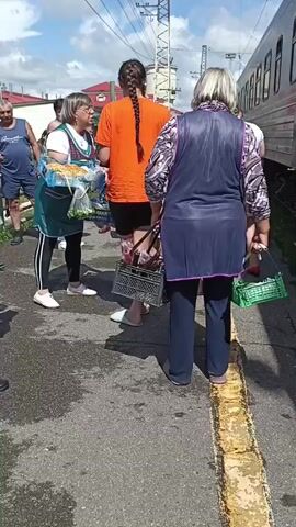 A group of people are standing on a wet train platform. A person wearing an orange shirt and black shorts with a braid is standing in the center, holding a phone to their ear. To their left, a woman wearing a green apron over a white shirt and black leggings is holding a package of produce. To the right of the person in orange, a woman wearing a dark blue apron over a patterned shirt and dark pants is holding a black basket. Other people are visible in the background, some near a train car. The platform has yellow markings along the edge. The sky is cloudy.