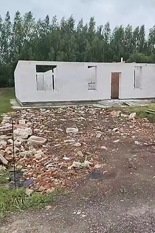 The media file shows a construction site for a new building, possibly a house, in a rural or semi-rural setting. The structure is partially built, with white walls and open doorways and windows. In the foreground, there is a pile of rubble, including bricks and stones, indicating demolition or debris from construction. The ground is a mix of dirt and grass. Behind the building, a dense line of tall green trees forms a backdrop, with a few scattered trees closer to the building. The sky is overcast with grey clouds, suggesting a possibly cool or damp day. There are no people visible in the frame. The overall impression is one of ongoing construction in a natural environment. The location is identified as Russkiye Dali, Russia.