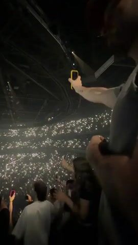 The video shows a concert audience from a close-up perspective, focusing on a person in the foreground holding up a phone with a yellow case. The background reveals a vast, dark arena filled with thousands of people, each holding up their phone, creating a sea of white lights. The camera is slightly shaky, suggesting it is being held by someone actively participating in the event. The overall atmosphere is that of a live music performance, with the crowd illuminated by their phone screens.