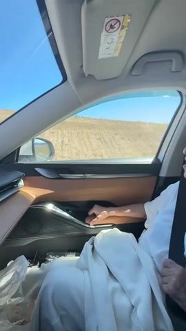 A woman in a white shirt and headscarf, wearing glasses and a seatbelt, sits in the passenger seat of a car. She is looking out the window at a dry, hilly landscape with plowed fields. The sun is shining, and the sky is blue. The interior of the car is visible, including the dashboard, door panel, and a sun visor with an airbag warning sticker. The woman's hand is resting on the door handle.