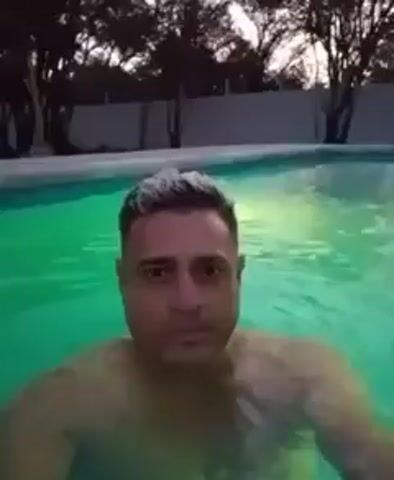 A man is in a swimming pool, holding the camera in front of him. The pool water is illuminated with green light. The man appears to be treading water, with his face and upper body visible. He has short hair, with the front section appearing lighter in color. Behind the pool, there is a white fence and some trees, with the sky visible above.