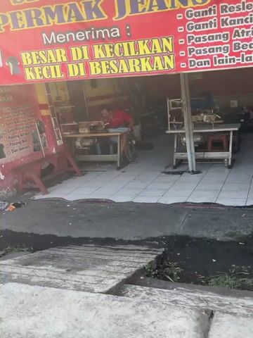 The video shows the exterior of a tailor shop. A red banner with yellow lettering hangs above the storefront, advertising "PERMAK JEANS" and listing services such as "Ganti Resleting" (zipper replacement), "Ganti Kancing" (button replacement), "Pasang Atrium" (patch application), "Pasang Belt" (belt loop repair), and "Dan lain-lain" (and more). Below the banner, a smaller yellow sign says "Menerima: BESAR DI KECILKAN KECIL DI BESARKAN" (Accepts: Big to Small, Small to Big). Inside the shop, a person wearing a red shirt is seated at a sewing machine. Another sewing machine is visible on a table to the right. The environment appears to be an outdoor street scene with a tiled floor inside the shop. A concrete structure, possibly a sidewalk or drainage system, is in the foreground.