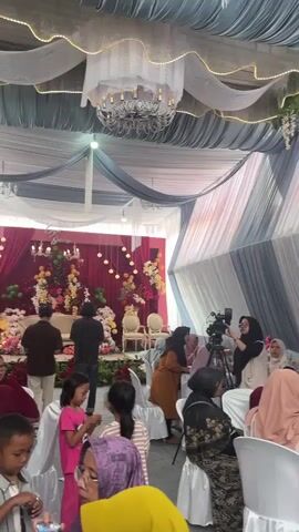 A wedding reception is underway under a draped tent. People are seated at tables, some eating and some watching performers. A stage with musicians is visible in the background, featuring a keyboard player and a singer. A camera operator is filming the event. The decor includes floral arrangements and draped fabric.