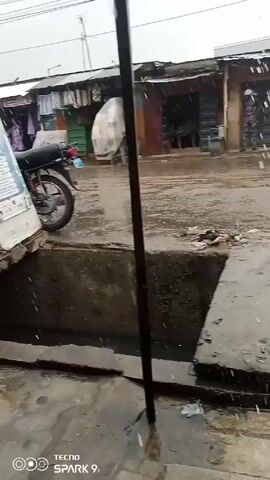 The video shows a rainy street scene in what appears to be a marketplace. The ground is wet and there are puddles. A sign for "POS available here" is visible near a concrete structure with a drain. A motorcycle is parked nearby. People are walking in the distance. The buildings in the background look like shops and residences. The weather is overcast and it's raining.