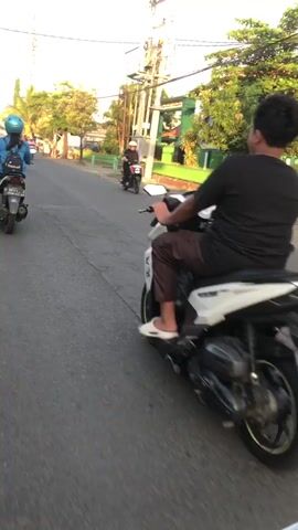 The video shows a group of people riding motorcycles on a paved road. The camera is mounted on a motorcycle and follows the riders. The first rider in the foreground is wearing a black t-shirt and brown pants. The second rider is wearing a pink shirt. The third rider is wearing a blue jacket. The fourth rider is wearing a white helmet. The road is lined with trees and buildings. The sky is clear and the sun is shining.