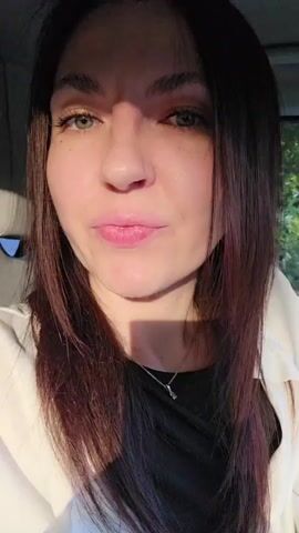 A woman with long, dark hair and green eyes is speaking to the camera. She is wearing a black top and a white jacket. The video appears to be filmed in a car, with trees visible in the background. The woman smiles and blows a kiss at the camera. The lighting suggests it is daytime, possibly early morning or late afternoon.
