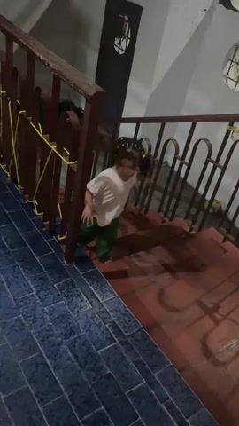 The video shows a toddler walking down a set of stairs inside a building. The toddler has curly brown hair tied back in pigtails adorned with small yellow flower clips. She wears a white t-shirt and green pants, and is barefoot. She moves slowly down the stairs, holding onto the railing for support. The railing is brown and connected with yellow rope. The stairs have red brick-like tiles, while the landing features blue tile. In the background, there is a white wall and a dark door with a decorative window. The toddler stops on the landing, looks at the camera, and talks. A voice responds, and the toddler continues to look at the camera. The lighting is dim, suggesting it is either night time or an interior space with limited light.