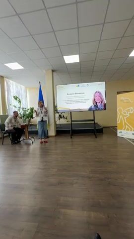 The video shows a presentation taking place in a well-lit room with a wooden floor and a tiled ceiling. A large screen displays presentation slides, and a woman is standing in front of it, speaking and gesturing. A man is seated to her left, appearing to listen. The presentation seems to be about community and public engagement, as suggested by the visible text on the screen which mentions "Миронівська" (likely a place name, referring to Myronivka), "громадська" (public/community), and "діяльність" (activity). The woman is wearing traditional Ukrainian clothing, specifically an embroidered shirt and a long denim skirt, indicating a cultural context. The screen displays the name "Богдана Безчастна" and a quote in Ukrainian that translates to "The strength of unity of the community in the protection of their land and future." A Ukrainian flag is visible in the background. The atmosphere appears formal and informative, likely a conference, seminar, or public meeting. The room is decorated with plants, and natural light streams in from large windows with white curtains, suggesting it is daytime.