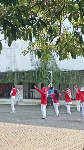 A group of people are engaged in an outdoor activity, possibly a dance or exercise routine, in a paved area. They are all wearing red tops and white bottoms, with several women also wearing headscarves. The individuals are positioned in a line or loosely spaced formation and appear to be following instructions from someone who is out of frame to the right. The background features a building with a white facade covered in graffiti and an abundance of green hanging plants. A tree with large leaves shades the scene from above. A metal structure, possibly a speaker stand or a stage support, is visible behind the group. The overall atmosphere is one of organized physical activity in a semi-urban outdoor setting.