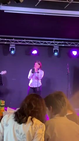 A young woman with short, dark hair stands on a stage, holding a microphone and singing. She wears a light-colored collared shirt and dark pants. She is illuminated by purple stage lights, and there is a guitar on a stand to her left. In the foreground, the backs of three people's heads are visible, suggesting they are an audience watching the performance. The environment appears to be an indoor venue with a dark background and exposed ceiling trusses with lighting equipment.