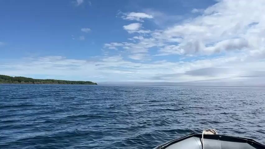 A view from the front of an inflatable boat sailing on the ocean. The water is choppy and blue, with white caps. The sky is blue with scattered white clouds. In the distance, there is a landmass with trees. The boat is moving towards the land. The camera is pointed forward, showing the bow of the boat and the vast expanse of the ocean.