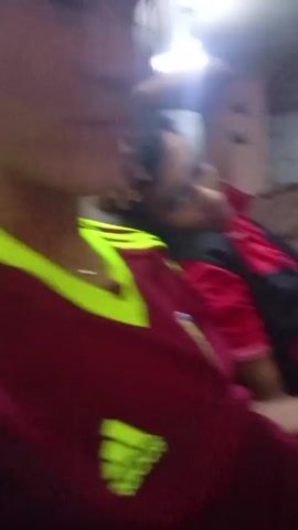 Two young men are in a room, likely a living room. The camera is held by one of the men, who is wearing a maroon Adidas soccer jersey with neon yellow accents. The other man is reclining on a brown couch, wearing a red shirt with a black vest. He is looking directly at the camera. The room has a slightly cluttered appearance, with various items visible in the background, including what appears to be a wall decoration. The lighting in the room is somewhat bright. The camera holder starts speaking.