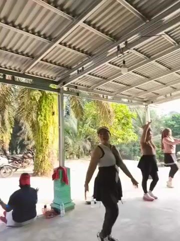 The video shows an outdoor exercise class under a covered patio. In the foreground, a woman in black leggings and a white and black top is performing an exercise with her arms raised. To her left, a woman in a pink long-sleeved shirt and pink pants is also exercising. Further to the left, a person in a hijab and a black skirt is visible. In the background, a person in a red head covering and dark clothing is sitting down. To the right, another person in a pink shirt and pink pants is exercising. The background consists of palm trees and foliage, with a motorcycle parked to the left. The overall setting appears to be a rural or semi-rural area. The participants are all in motion, engaging in synchronized movements.