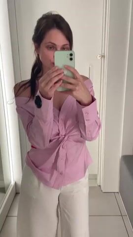 A woman stands in what appears to be a fitting room, taking a selfie with a smartphone. She is wearing a pink and white striped off-the-shoulder shirt, tied at the waist, and loose white pants. She has her hair pulled back in a ponytail. The room has bright lighting, a full-length mirror to her left, and a door to her right. She makes a few different poses for the camera, including puckering her lips and adjusting her shirt.