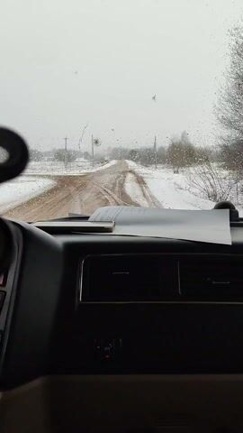 A hand holding a black plastic triple mount is filmed inside a car. The camera is at dashboard level and shows the windshield in the background. It is snowing, and the road can be seen in the background. The hand holding the mount rotates it slightly. A piece of paper is on the dashboard. A straw or tool rests on top of the paper. Compressed air is used to clean the mount.