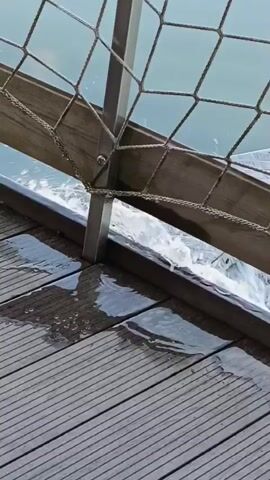The video shows a close-up shot of a wooden deck with a metal railing and a net. Water is flowing over the edge of the deck and splashing onto the wooden planks. The water appears to be moving rapidly and is creating ripples and splashes. The overall impression is one of movement and the force of water.