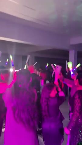 A crowd of people is dancing indoors, most waving long, white, glowing, color-changing light wands over their heads. The room has a low, white ceiling with recessed lighting. The lighting above is provided by changing colored lights. People are dressed in a variety of formal attire, including dresses, blouses, suits, and blazers. The floor is visible in places and appears to be light-colored tile. Some columns are along the back wall.