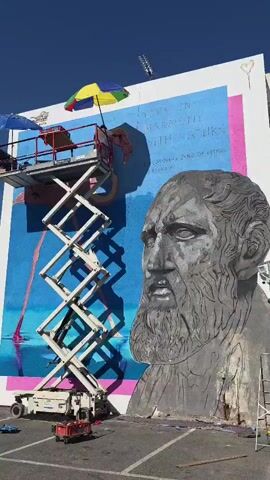 This video captures a mural being painted on an outdoor wall. The mural features a large, grey bust of an ancient Greek philosopher with a long beard and furrowed brow. To the left of the philosopher's head, a vibrant blue section of the mural is visible, with pink and yellow text overlaid. The text reads: "LIVE IN HARMONY WITH NATURE" and beneath it, "PHILOSOPHER ZENO OF KITIUM."

A scissor lift is positioned in front of the mural, elevated to reach a section of the artwork. On the platform of the lift, a person wearing a white shirt and a dark cap is actively painting. They are working on a detail of the mural, which appears to be a pink and yellow feathered creature, possibly a flamingo or a stylized bird, with a curled, yellow form beneath it. A colorful umbrella, with panels of yellow, green, blue, and red, is attached to the lift, providing shade for the artist. Another smaller blue umbrella is also visible on the platform.

The scene appears to be set outdoors, suggested by the clear blue sky and the bright sunlight casting shadows. The ground beneath the lift is a paved surface, possibly a parking lot or street. The time of day is clearly daytime, with strong sunlight. The overall impression is one of artistic creation in progress, with the artist diligently working on the large-scale mural. The setting in Larnaca, Cyprus, adds a geographical context to the scene.