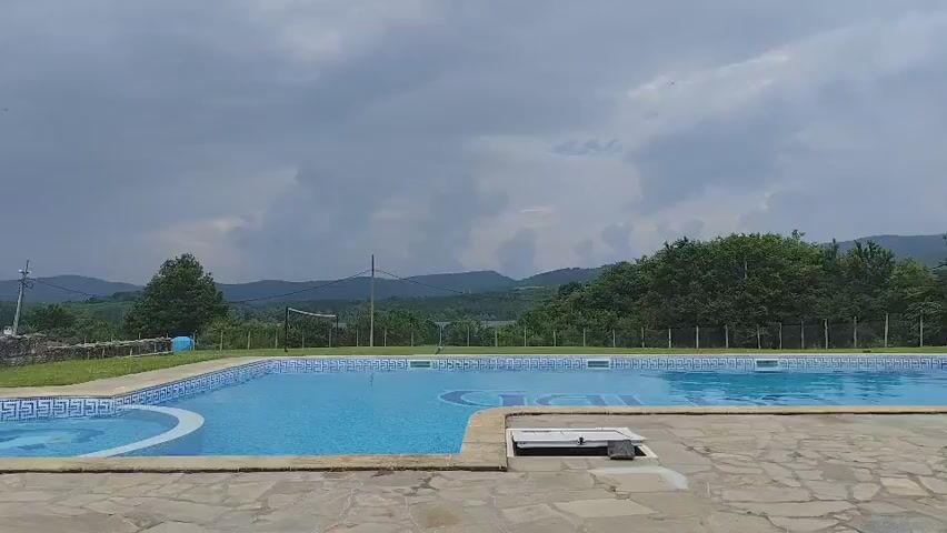 A wide shot shows an outdoor swimming pool on a cloudy day. The pool is blue and has a light blue tiled border with geometric patterns. The pool is surrounded by light brown flagstones and a strip of green grass. In the background, there is a panoramic view of lush green hills and a dark gray, overcast sky. Some trees are visible on the left and right sides of the frame. The water in the pool is still, reflecting the cloudy sky.