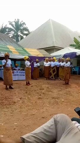 A group of women are performing outdoors in front of a tent with the words "ST. ANTHONY OF PADUA INDU/AMANU STATION" displayed. They are dressed in matching patterned skirts, white blouses, and yellow headscarves. Two women stand forward, seemingly leading the performance with microphones. Behind them, other women are standing and singing. Behind the performers is a table with people seated, a drum set, and chairs. The ground is reddish dirt with small stones. The video is shot from a low angle, showing a person lying down in the foreground. The setting appears to be a community gathering.