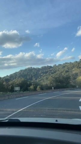 The video shows a view from inside a car driving on a winding road through a hilly, wooded area. The road is asphalt with white lane markings and a concrete barrier on one side. A silver car drives towards the camera on the opposite lane. The hillside is covered in dense green trees. The sky is blue with scattered white clouds.