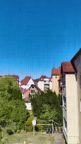 This is a video of a suburban backyard in the daytime. The camera pans from right to left, showing a white car parked on the street, a white tarp covering something in the backyard, various trees and shrubs, two trash bins, a satellite dish, and several houses with red roofs in the background. The sky is a clear blue. There are no people visible in the video.