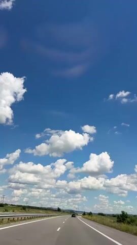 The video is a first-person perspective shot from a vehicle moving along a highway. The sky is a vivid blue with scattered white cumulus clouds. A few larger, darker clouds are visible in the distance. The highway has two lanes in each direction, separated by a white dashed line. A metal guardrail runs along the left side of the road. Fields of green grass and vegetation line both sides of the highway. A dark-colored car is visible in the distance, traveling in the same direction. The overall impression is a bright, sunny day on an open road.