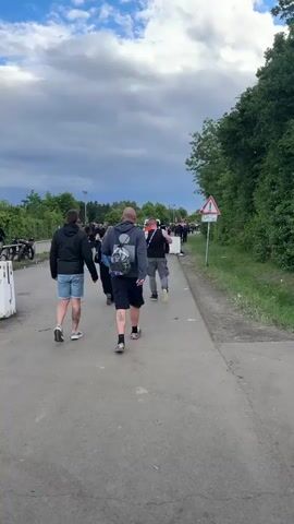 A wide shot shows a crowd of people walking on a paved road toward a concert venue. The road is bordered by a metal fence on the left and a patch of grass with dense trees on the right. There's a white sign with a red border and a black exclamation mark on the right. The sky is partly cloudy, with blue sky visible at the top and darker clouds closer to the horizon.