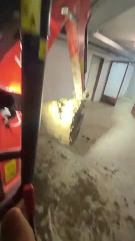 A front loader dumps a scoop of dirt onto the floor of a concrete room. The front loader is red and black, with yellow warning stickers. It faces a white cinderblock wall. A second piece of red and white heavy machinery is on tracks, and positioned to receive the contents of the front loader's bucket. The room is lit by overhead fluorescent lights. The camera is hand-held and relatively stable.