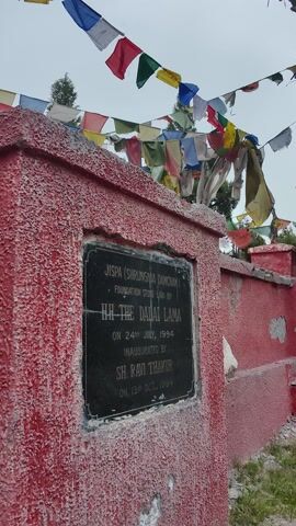 This media file shows a memorial plaque on a red, weathered stone structure. The plaque is black and inscribed with white text. The text reads:

"JISPA (SHRUNGMA DAMCHAN)
FOUNDATION STONE LAID BY
H.H. THE DALAI LAMA
ON 24TH JULY, 1994
INAUGURATED BY
SH. RAVI THAKUR
ON 13TH OCT, 1994"

The memorial is decorated with numerous colorful Buddhist prayer flags strung across the foreground and upper left of the frame, fluttering in the wind. The background shows a cloudy or overcast sky and some greenery, suggesting an outdoor setting. The stone structure appears to be outdoors, possibly on a hill or elevated area. The weather appears to be cloudy or hazy. The overall mood is respectful and commemorative. The setting is likely a significant site in Jispa, India, given the mention of the Dalai Lama.