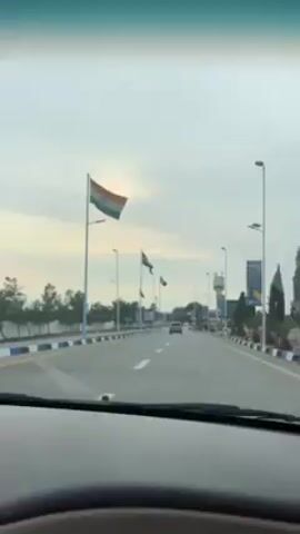 The video shows a car's perspective driving down a road lined with flagpoles. The flags include those of India, Jordan, and France. The road is paved, with white lane markings and blue and white curbs.  Streetlights line the road, and there is some greenery visible. The sky is overcast, and the sun is visible as a bright circle behind the clouds. There is a single car in the distance ahead. A billboard is visible to the right of the road.  The dashboard of the car is visible at the bottom of the frame.  Music plays in the background.