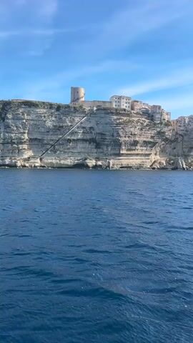The video shows a sea view of a coastal city perched atop a tall, rugged cliff face. The sky is a clear, bright blue with a few scattered clouds. The sea is a deep blue, with gentle waves rippling across its surface. The cliff is composed of layered rock formations, with buildings sitting precariously on the edge. There is a tower on top of the cliff on the left side of the frame. A long, dark object, perhaps a staircase or ramp, stretches down the side of the cliff.