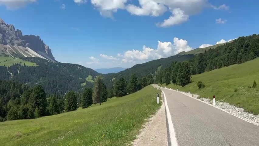 A group of motorcyclists ride along a winding mountain road. The scene is set in a picturesque mountain landscape with lush green hillsides dotted with evergreen trees. In the background, a majestic, rocky mountain peak rises against a clear blue sky filled with fluffy white clouds. The road is paved and has a white dividing line.

There are at least three motorcyclists visible. The first motorcycle is in the foreground, with the rider wearing a red and white helmet and black and white riding gear. The other two riders are further up the road, also on motorcycles. The time of day appears to be daytime, with bright sunlight casting shadows, suggesting a sunny and pleasant day. The overall atmosphere is one of adventure and scenic travel. The location cues, such as the distinctive mountain formation and the Italian road signs (though not clearly legible), point to the Dolomites region of Italy.