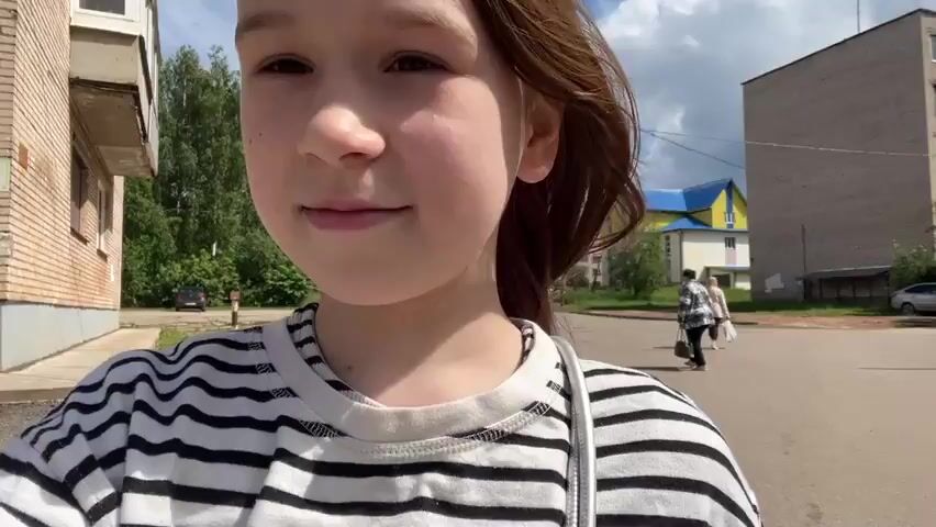 The video shows a close-up of a young person with light brown hair and fair skin, wearing a black and white striped t-shirt. The person is outdoors, with a blurred background of buildings, trees, and a blue sky with clouds. The person's right hand comes into frame and touches their hair. In the background, two people are walking away from the camera down a paved path. To the right, several green trash bins are visible. The person is facing the camera and looking slightly to the left of the lens. The overall impression is a casual, handheld shot, likely a selfie or a vlog-style recording.