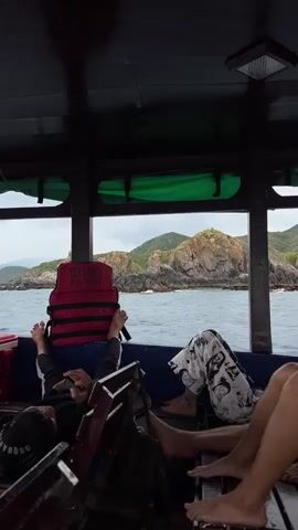 Inside a covered boat, three people are relaxing on benches with their feet up facing the open window. One person is lying down, looking at their phone. In front of them is a red life vest with "YEN SAO 01" and "KH 0315" printed on it. Outside the window, there is a rocky island covered in green vegetation. The sea is visible in the foreground. The boat interior is dark and made of wood.