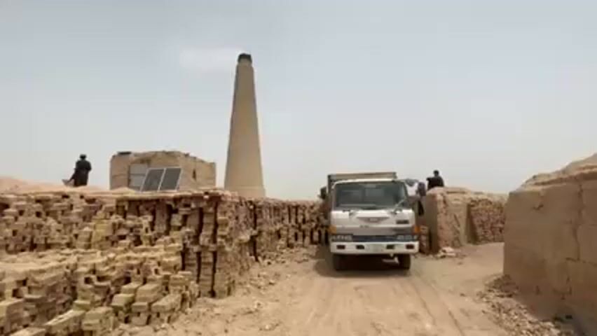 The video shows a truck parked in front of a brick kiln. There is a tall chimney behind the truck, with smoke rising from it. To the left, a pile of unfired bricks are stacked next to a small building. A solar panel is mounted on the building. Two men can be seen. One man is standing on the small building to the left. Another man stands next to a brick structure on the right. The ground is dirt, and the sky is hazy.