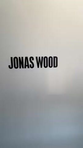 The video begins with the text "JONAS WOOD" displayed on a white background. The camera then moves through a hallway with white walls on the left and windows on the right. Several people are gathered at the end of the hallway. The hallway has a smooth, gray floor.
